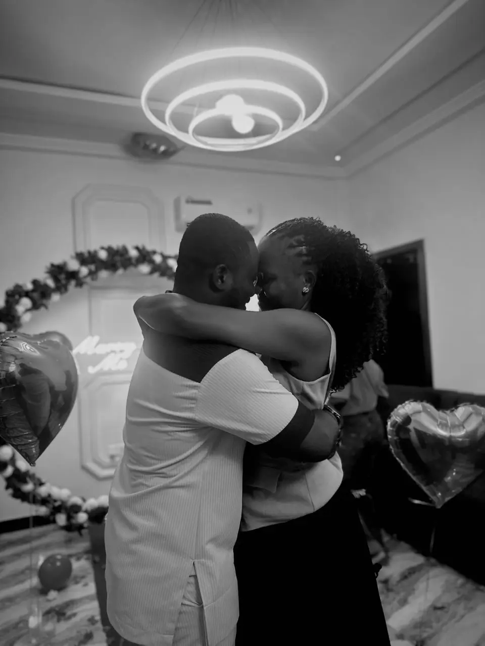 Noblet and Nancy embrace beneath a neon Marry Me sign, in black and white
